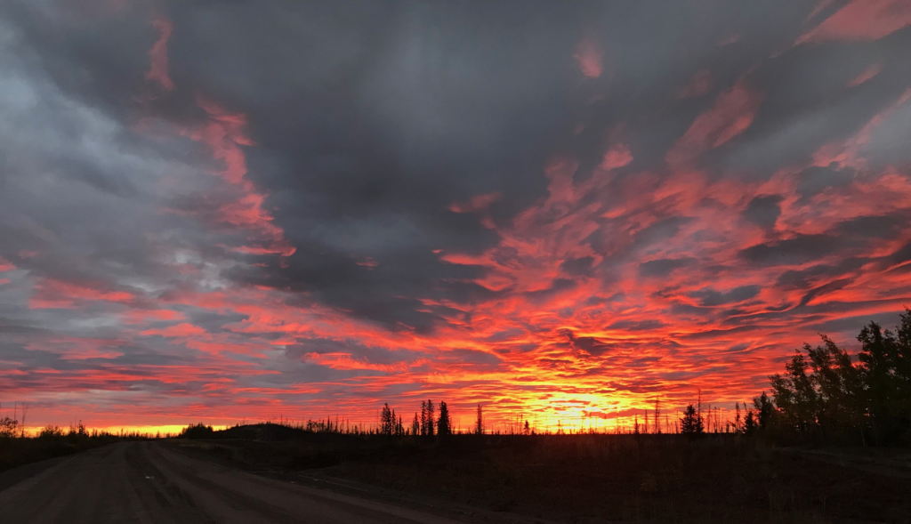 sunrise against overhead clouds above dark road with trees on the skyline